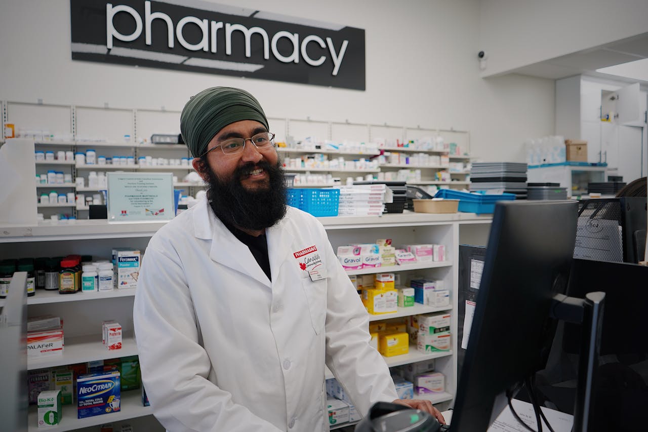 A pharmacist working at a computer terminal inside a pharmacy, with shelves of medication in the background.