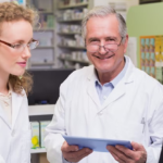 Two smiling pharmacists using pharmacy retail software on a computer and tablet