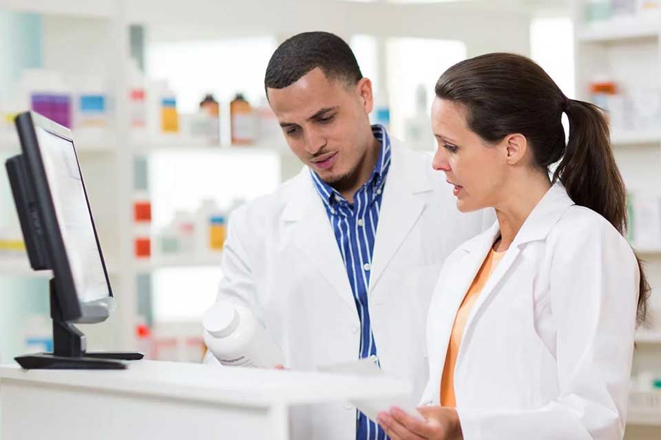 Two people in lab coats and a pharmacy setting discussing in front of a computer.
