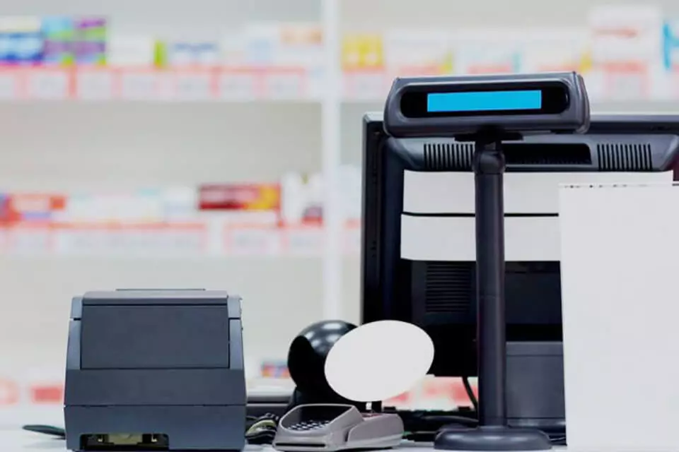 A receipt printer, barcode scanner, customer display, and computer monitor with shelves of medications in the background