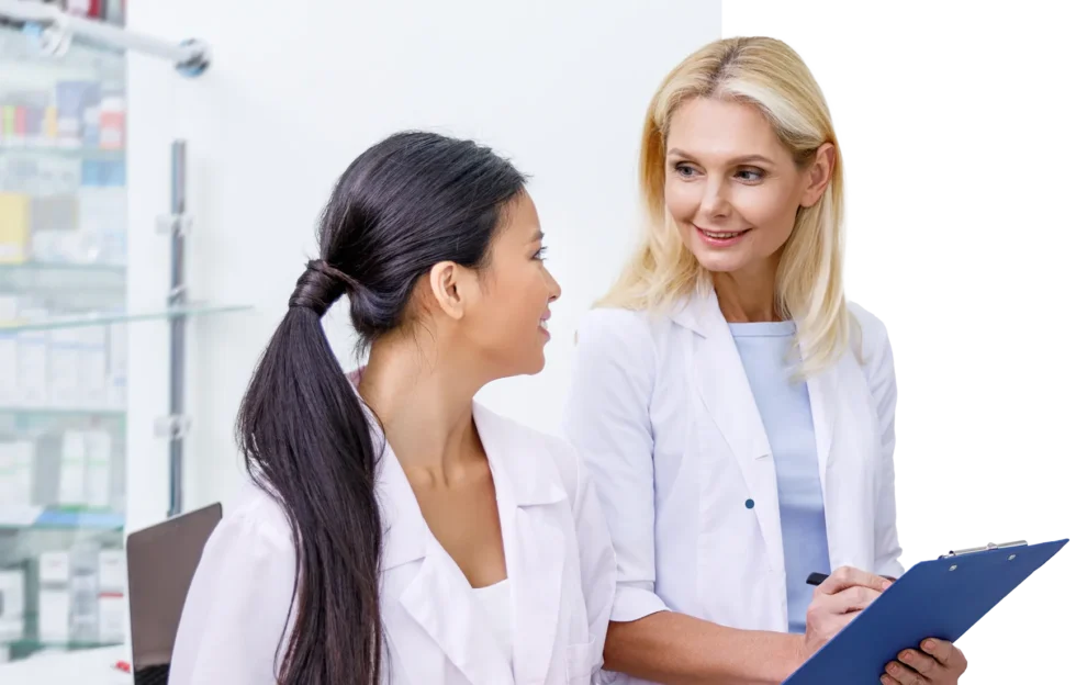 Two pharmacists in white coats have a friendly discussion while reviewing notes on a clipboard.