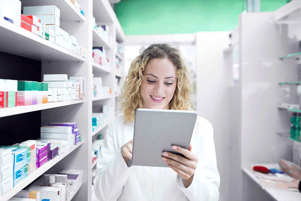 A pharmacist stands between stocked shelves, smiling while using a tablet