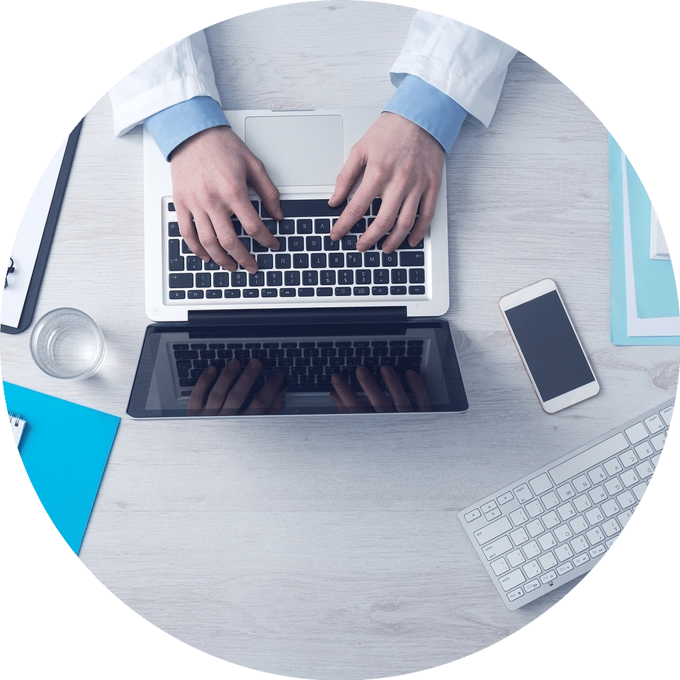 A top-down image showing a person’s hands resting on a laptop with a phone, glass and a keyboard on the desk too.