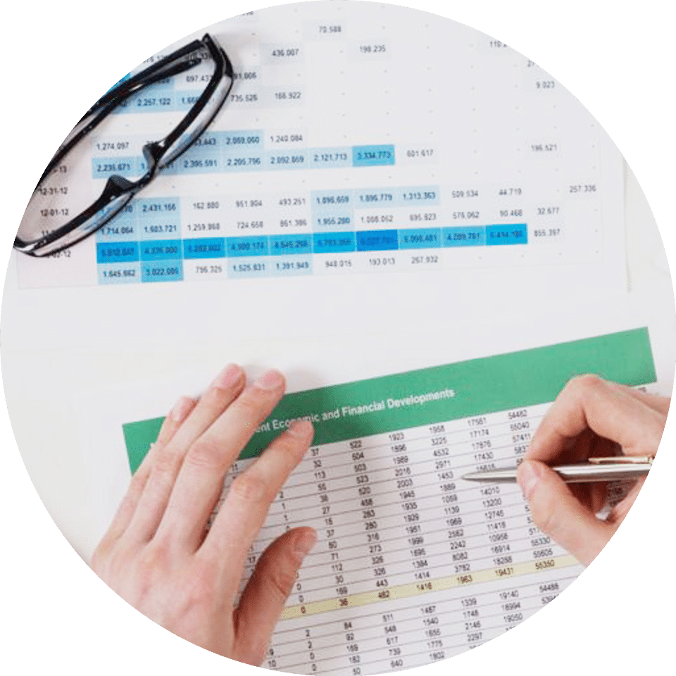 A picture showing several printed spreadsheets and graphs with a laptop to the side and a person’s hands.
