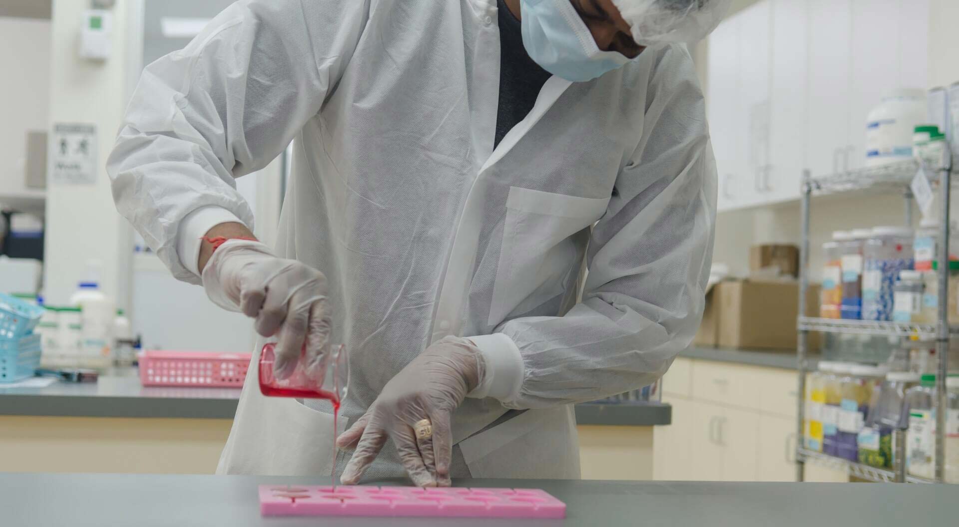 A photo of a pharmacy technician wearing protective gear while pouring a red liquid into a silicone mold in a compounding lab.