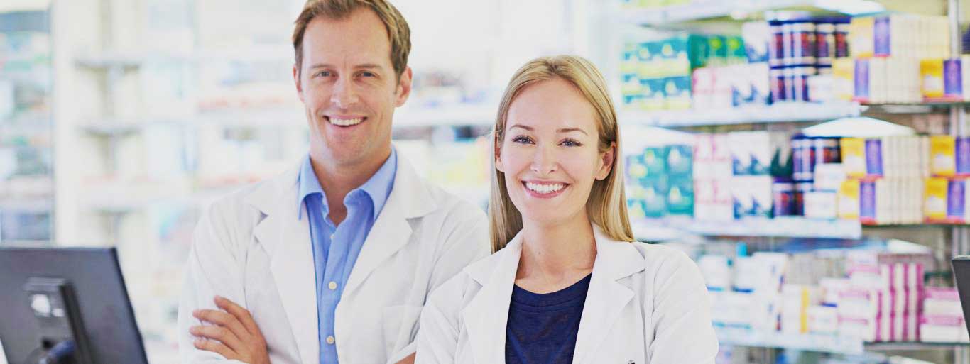 A man and woman dressed in lab coats in a pharmacy setting as they pose for the camera