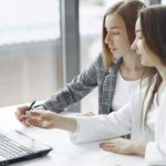 A pharmacist and her assistant discuss pharmacy management systems in front of a laptop