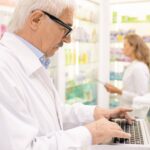 A white-haired pharmacist with glasses looking up information with pharmacy software on a laptop in a pharmacy